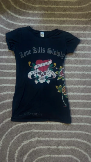 Svart Ed Hardy t-shirt med tryck - Cool svart t-shirt från Ed Hardy med korta ärmar och färgstarkt tryck. Framsidan har motiv med dödskalle, hjärta och texten 'Love Kills Slowly', samt blommor i rött, gult och grönt. Baksidan har en stor lila ros och Ed Hardy-logga. Materialet är mjuk bomull.