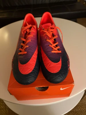 Nike Hypervenom Phantom II SG fotbollsskor - Nike Hypervenom Phantom II SG fotbollsskor i storlek 45. Snygg design med röd, lila och svart färg, orange snörning och stora Nike-loggor. Skorna har rund tå, platt sula och metall-dobbar för bra grepp på gräs. Tillverkade i syntetmaterial för lätt känsla på planen. Aldrig använda 