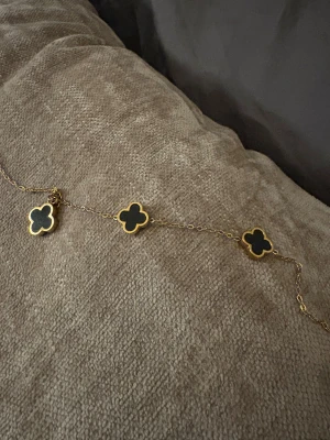 Guldigt halsband med svarta klöver - Snyggt halsband med tunna guldfärgade länkar och tre svarta fyrklöverformade hängen med guldkant. Perfekt för dig som gillar stilrena och unika detaljer. Klöverhänget ger en lyxig och modern vibe.