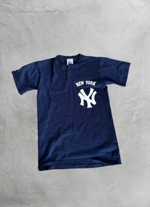 New York Yankees t-shirt från Majestic - Mörkblå New York Yankees t-shirt från Majestic med vit logga och text på bröstet. Klassisk rak passform, korta ärmar och två knappar vid halsen. Tillverkad i mjuk bomull, perfekt för dig som gillar sportig och avslappnad stil. Storlek S/M.