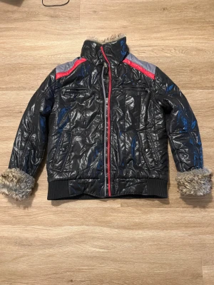 Svart pufferjacka med pälsfoder Mondo - Svart pufferjacka från Mondo Casual med glansig yta, röda och grå detaljer på axlarna och dragkedja framtill. Jackan har fuskpälsfoder och päls vid ärmsluten, samt flera fickor med dragkedja. Perfekt för kalla dagar med sin fluffiga insida.