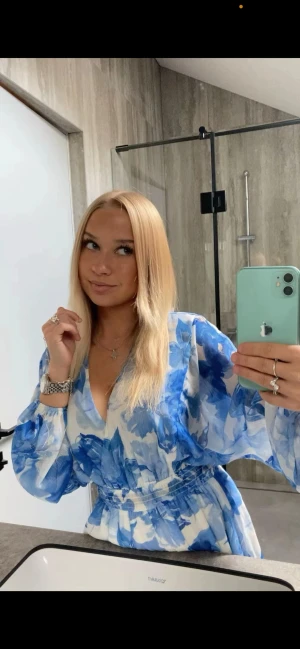 Blåvit blommig volangklänning - Superfin kort  volangklänning i blått och vitt med blommigt mönster. Klänningen har v-ringning, långa puffärmar och markerad midja med resår. Tunt, luftigt material som passar perfekt till varmare dagar. Snygg och trendig look!