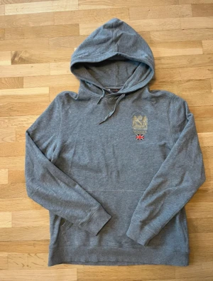 Grå tröja från Morris med luva och broderi - Snygg grå hoodie från Morris med guldbroderat emblem och en liten brittisk flagga på bröstet. Tröjan har huva med snörning och långa ärmar, perfekt för chill dagar. Mjuk bomullskvalitet och klassisk passform.
