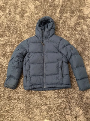 Blå Peak Performance dunjacka - Säljer en blå dunjacka från Peak Performance med huva och diskret logga på ärmen. Jackan har quiltad design, dragkedja framtill och två stora fickor. Perfekt för kalla dagar och har en clean, sportig look som funkar till allt.                                           Priset är ej 100% satt kan gå ner.                                    Storlek är L men Passar XL stor i form är den,             Den är använd cirka 1 månad allt som allt.                  Produkten säljs ej längre i affär