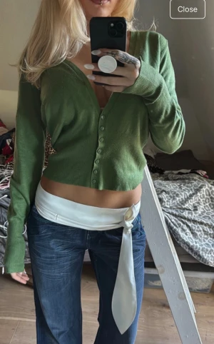 Grön croppad kofta med v-ringning - Snygg grön croppad kofta med v-ringning och knappar framtill. Tröjan har lång ärm och är gjord i ett mjukt stickat material som sitter loose. Perfekt för 90s look och för vintern. Det stör storlek 40 men sitter som en 38. Obs bilder är tagna av förra ägaren!!😚