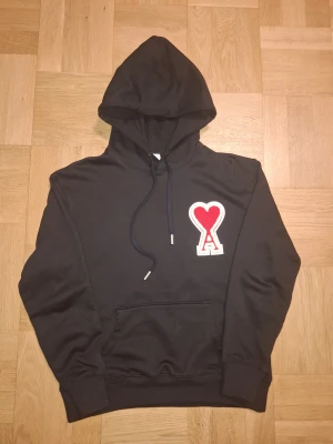 Ami Paris - Svart hoodie från Ami Alexandre Mattiussi med stor röd hjärtdetalj och bokstaven A på bröstet. Tröjan har huva med snörning, magficka och ribbade muddar. Tillverkad i mjuk bomull, perfekt för chill dagar och streetwear vibes. Perfekt för dig som har ett partner som börjar på A. Pris går att diskuteras!!!
