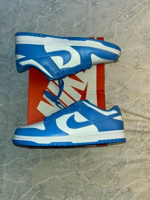 Nike Dunk Low blå och vita sneakers - Nike Dunk Low sneakers i fräsch blå och vit färg med klassisk design. Skorna har en låg siluett, rund tå och snörning. Tillverkade i skinn med perforerad tåbox för extra ventilation. Snyggt kontrasterande Nike-logga på sidan och tunga. Storlek 41