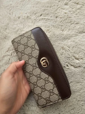 Plånbok med GG-mönster - Snygg plånbok med klassiskt GG-mönster i beige och brunt. Plånboken har detaljer i mörkbrunt skinn, guldfärgad logga och dragkedja runt om. Insidan har flera fack för kort och mynt, perfekt för att hålla ordning på allt.