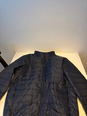 Mörkblå pufferjacka med dragkedja - Snygg mörkblå pufferjacka med ståkrage och hel dragkedja framtill. Jackan har ett quiltat mönster och är långärmad, perfekt för kyliga dagar. Materialet är syntet och jackan har en clean och modern look. Helt ny.