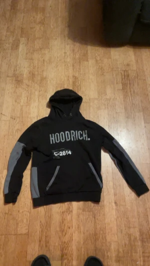 Svart och grå hoodie från Hoodrich - Säljer en svart hoodie från Hoodrich med grå detaljer på ärmar och fickor. Stor HOODRICH-text på bröstet och tryck med C-2814. Tröjan har huva med snörning och en klassisk känguruficka framtill. Perfekt för dig som gillar streetwear.