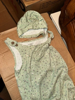 Grön blommig byxdress från Primark Baby Grön Blommig Sparkdräkt med Mössa - Söt grön byxdress med små blommor från Primark Baby, passar barn 12-18 månader. Mjuk bomullskvalitet, ribbad struktur och praktiska knappar på axlarna och längs benen. Ficka framtill och matchande mössa med rosett ingår.Söt grön blommig sparkdräkt med en liten ficka framtill och matchande mössa. Sparkdräkten har knappar på axlarna.