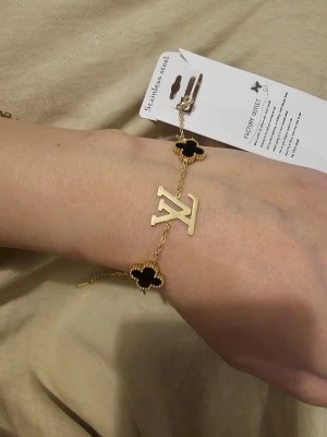  guldarmband - Snyggt armband i guldtonad metall med svarta fyrklöverformade detaljer och LV-logga. Kedjan är tunn och elegant, och armbandet har även små guldfärgade symboler som ger en lyxig känsla. Perfekt accessoar för dig som gillar statement-smycken.