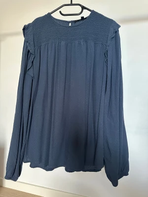 Blus från Kappahk - Säljer en mörkblå blus med långärmade och löst sittande ärmar. Blusen har smock upptill, volangdetaljer vid axlarna och ett diskret mönster i tyget. Rund halsringning och knapp i nacken  Obs: Alla plagg säljs endast fram till 28/12