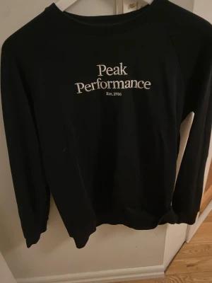 Svart sweatshirt Peak Performance - Svart sweatshirt från Peak Performance med vit broderad logga på bröstet. Tröjan har rund hals, långa ärmar och ribbade muddar vid ärmslut och nederkant. Perfekt för dig som gillar stilren och enkel design.