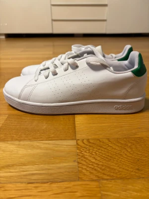 Adidas Stan Smith vita sneakers storlek 38,5 - Fräscha vita Adidas Stan Smith sneakers med gröna detaljer på hälen och klassisk snörning. Skorna har perforerade sidor, rund tå och platt sula. Tillverkade i syntetmaterial med mjukt foder och stilren design. Perfekta för dig som gillar cleana och tidlösa sneakers.