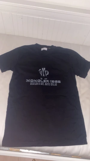 Svart Moncler 1952 t-shirt i bomull - Svart t-shirt från Moncler med tryck 'Moncler 1952 Genius & Est. 2018 Milan' i silver på bröstet. Klassisk rund hals och korta ärmar. Materialet är mjuk bomull och passformen är normal. Perfekt för dig som gillar exklusiva streetwear-vibes.