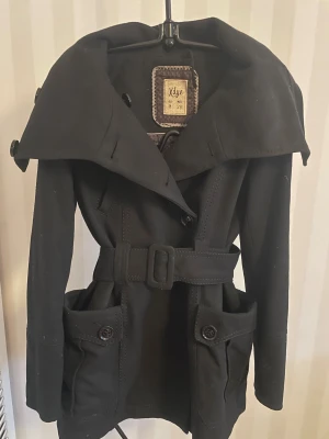 Svart trenchcoat från Xdye med bälte - Snygg svart trenchcoat från Xdye med bred krage, stora fickor med knappar och ett matchande bälte i midjan. Jackan har långa ärmar och stilrena detaljer som ger en cool och modern look. Perfekt för dig som gillar klassisk stil med edge.