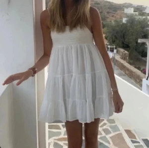 Vit kortklänning med volanger - Supersöt vit kortklänning med tunna axelband och volanger i flera lager. Klänningen har en luftig passform och är tillverkad i ett lätt material som ger en skön känsla. Perfekt för varma dagar och ger en fräsch och ungdomlig vibe.