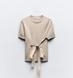 Beige stickad topp med knyt från Zara - Superfin beige stickad topp från Zara med korta ärmar och rund hals. Toppen har en snygg knytdetalj i midjan som ger en unik look. Perfekt att styla med jeans eller kjol för en trendig outfit.