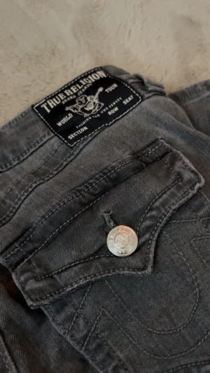 True Religion grå bootcut jeans - Snygga grå jeans från True Religion med bootcut passform. Jeansen har klassiska fickor med lock och silverfärgade knappar med logga. Materialet är jeans och de har tydliga sömmar och detaljer som ger en cool vibe.