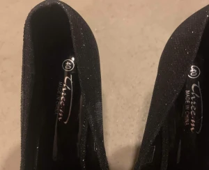 Svarta glittriga pumps med öppen tå - Säljer ett par svarta pumps från Three-in med glittrig yta och snygga utskurna blommönster. Skorna har hög klack och öppen tå, vilket ger en extra touch till outfiten. Perfekta för dig som vill sticka ut med elegans och stil.passar st /39