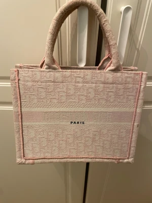 Handväska cd  - Säljer en lyxig handväska i ljusrosa och beige ton. Väskan har broderad logga, Paris-text och är tillverkad i bomull med strukturerat mönster. Den har två rejäla handtag och en boxig, fyrkantig form som rymmer mycket.