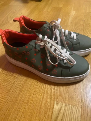 MCM sneakers i grönt och rött - Snygga sneakers från MCM i matt grön färg med röda detaljer och logomönster. Skorna har vit platt sula, vita snören och röd insida. Tillverkade i skinn med klassisk rund tå och låg modell. Perfekta för dig som vill sticka ut med exklusiva detaljer.