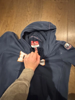 Mörkblå hoodie från Parajumpers - Snygg mörkblå hoodie från Parajumpers med stort PJS-tryck i vitt och orange på bröstet. Tröjan har huva, lång ärm och en klassisk känguruficka framtill. Patch med logga på ena ärmen och mjukt fleece-material som gör den extra skön.