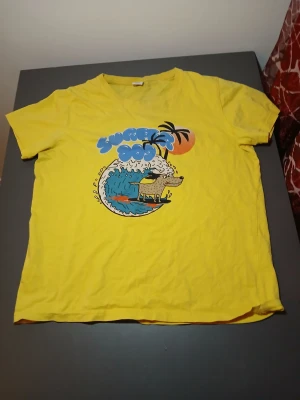 Gul Surfer Dog t-shirt med tryck - Gul t-shirt med coolt Surfer Dog-tryck framtill, där en hund surfar på en våg med palmer och solnedgång i bakgrunden. T-shirten har korta ärmar och v-ringad hals. Perfekt för dig som gillar färgglada och roliga motiv.