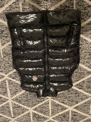 Svart dunväst från Moncler - Snygg svart dunväst från Moncler med glansig finish och klassisk logga på framsidan. Västen har två fickor med dragkedja och quiltad design. Perfekt för lager-på-lager och streetstyle. Stängs med dragkedja framtill.