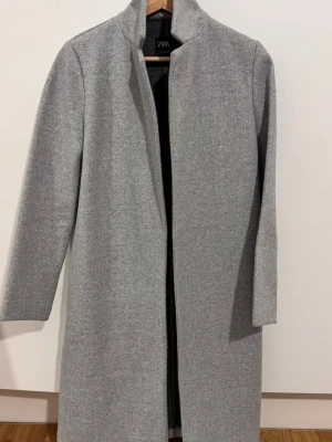 Grå kappa från ZARA med ståkrage - Snygg grå kappa från ZARA med stilren ståkrage och raka linjer. Jackan är lång, har en minimalistisk design utan synliga knappar eller fickor och är tillverkad i ett mjukt ullblandat material. Perfekt för dig som gillar clean och modern look.