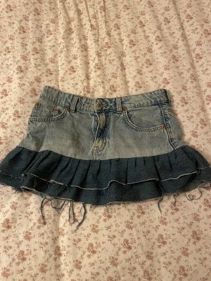 Blå jeanskjol med volang från H&M Divided - Säljer en kort jeanskjol från H&M Divided i ljusblå denim med mörkare blå volang nedtill. Kjolen har klassiska fickor fram och bak, bälteshällor och råa kanter med slitna detaljer. Perfekt för dig som gillar en edgy och trendig look. Jag har sytt den själv!