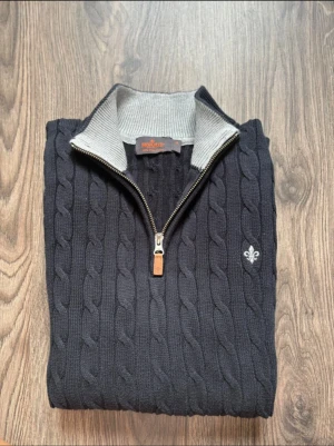 Blå kabelstickad tröja från Morris - Snygg marinblå kabelstickad tröja från Morris med halv dragkedja och ribbad grå krage. Tröjan är i 100% Pima cotton och har långärmade armar med förstärkta armbågslappar. Snygg broderad logga på bröstet och coolt stickat mönster. Perfekt för dig som gillar klassisk stil. 