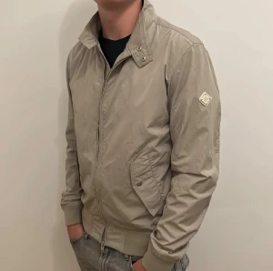 Beige vindjacka från J.Lindeberg - Snygg beige vindjacka från J.Lindeberg med ribbade muddar, ståkrage med tryckknappar och dragkedja framtill. Jackan har två fickor med lock och en diskret logotyp på vänster ärm. Perfekt för dig som gillar stilren och enkel design.