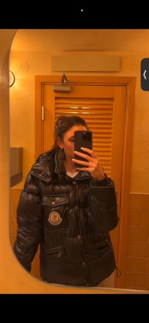 Svart Moncler pufferjacka med logga - Säljer en svart pufferjacka från Moncler med stor logga på bröstet. Jackan har hög krage, flera fickor med knappar och dragkedja, samt en glansig finish. Perfekt för dig som vill ha en varm och snygg jacka med street vibe.