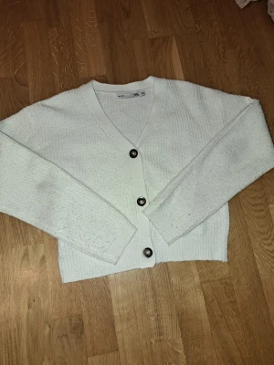 Vit stickad kofta från 157 - Mysig vit stickad kofta från 157 med v-ringning och tre stora knappar framtill. Koftan är lite noppring men inget man tänker på. Perfekt för lager-på-lager och chill dagar. Snyggt croppad passform som ger en modern vibe.