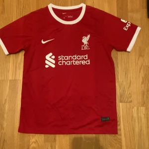 Liverpool 23/24 fotbollströja - Liverpool 23/24 matchtröja från Nike i rött med vita detaljer vid krage och ärmslut. Tröjan har klubbmärke och sponsortryck på bröstet samt Nike-logga. Tillverkad i lätt och ventilerande material, perfekt för fotboll eller supporterliv.