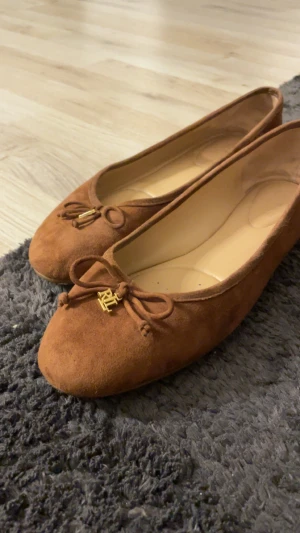 Bruna mocka ballerina Ralph Lauren - Snygga ballerina från Ralph Lauren i brun mocka med rund tå och dekorativ rosett framtill, prydd med guldfärgad RL-logga. Skorna har platt sula med gummidetaljer under hälen för extra grepp. Perfekt för dig som gillar klassisk och stilren design. Lite slitna under ifrån