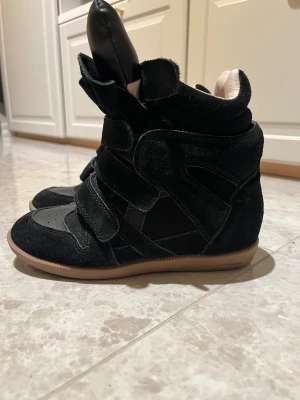 Isabel Marant liknande  svarta wedge sneakers - Isabel marant liknande wedge sneakers, mocka och skinn med beige insida och gummisula. Skorna har kardborreband, hög plös och cool, sportig design. Perfekta för dig som vill ha en edgy och trendig look med extra höjd. Använda en gång inga defekter 
