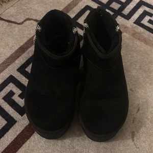 Svarta boots med mönstrad kant - Svarta boots i mocka med grov platt sula och rund tå. Skorna har en snygg vit och svart broderad detalj runt kanten upptill. Perfekta för dig som gillar stilrena och bekväma skor med lite extra edge.