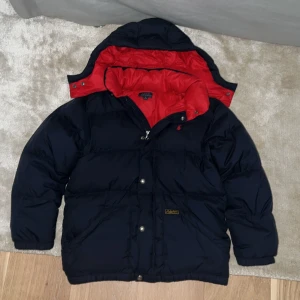 Blå pufferjacka från Polo Ralph Lauren - Säljer en blå pufferjacka från Polo Ralph Lauren med röd insida och blå utsida . Perfekt för kalla dagar under vintern och dig som gillar grisch stilen. Finns en liten defekt under luvan (bild 5) som ej ses när man använder den därför det låga priset!