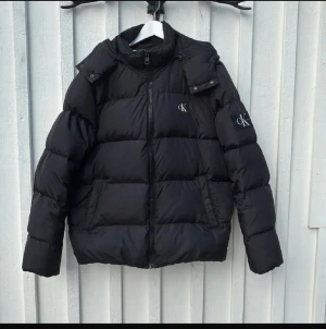 Svart pufferjacka Calvin Klein Jeans XL - Svart pufferjacka från Calvin Klein Jeans i storlek XL. Jackan har huva, dragkedja framtill och CK-logga på bröstet och ärmen. Den är vadderad och har en clean, modern look som passar perfekt när det är kallt ute.