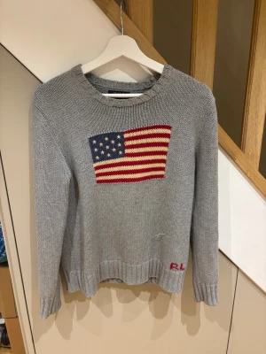 Grå stickad tröja Polo Ralph Lauren - Snygg grå stickad tröja från Polo Ralph Lauren med amerikansk flagga broderad på bröstet och RL-broderi vid nederkanten. Rund hals och långa ärmar, perfekt för dig som gillar klassisk stil med en twist.