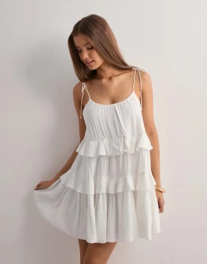 Vit volangklänning med öppen rygg - Supersöt vit kortklänning med tunna axelband som knyts och flera lager volanger. Klänningen har en djup öppen rygg med smal rem och är tillverkad i ett lätt, luftigt material. Perfekt för varma dagar och ger en fräsch och ungdomlig vibe. Säljer pga aldrig används, Orginalpris 399💝 använd 1-2 gånger ✌️