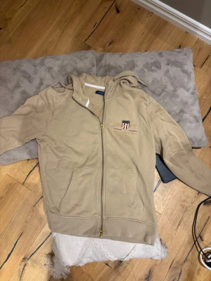 Beige hoodie från GANT med dragkedja - Snygg beige hoodie från GANT med broderad logga och emblem på bröstet. Tröjan har huva med snörning, praktisk dragkedja och fickor framtill. Perfekt för chill dagar och har en klassisk, clean look som funkar året runt. Passar folk i storlek s-m