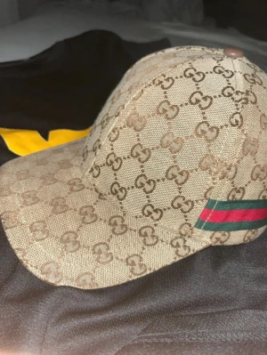 Beige Gucci keps med GG-mönster - Snygg keps från Gucci med klassiskt GG-monogram i brunt över hela kepsen. På sidan finns den ikoniska gröna och röda randen. Kepsen är tillverkad i bomull och polyester, med insida i svart tyg och Gucci-loggor. Perfekt för dig som vill ha en lyxig streetstyle.