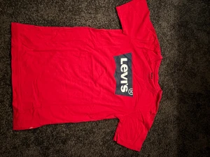 Röd Levi's t-shirt med logga - Snygg röd t-shirt från Levi's med klassisk stor logga i vitt och mörkblått på bröstet. T-shirten har rund halsringning och korta ärmar. Tillverkad i mjuk bomull som känns skön mot huden. Perfekt för en avslappnad och trendig look.