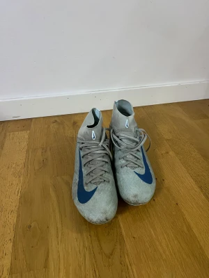 Nike Air Zoom grå/blå fotbollsskor - Säljer ett par Nike Air Zoom fotbollsskor i grått med blå detaljer och snörning. Skorna har en strumpliknande hög krage och platt sula med dobbar för gräsplan. Snyggt blått swoosh på sidan och grafiska linjer bak. Perfekt för dig som vill ha stil på planen.