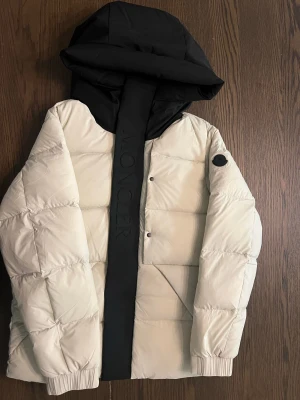 Moncler madeira  - Säljer en riktigt snygg beige Moncler dunjacka med stor svart huva och bred svart dragkedja med Moncler-logga. Jackan har puffig design, två sidofickor och svarta detaljer. Perfekt för kalla dagar och ger en clean, modern look. Helt ny finns i alla storlek.