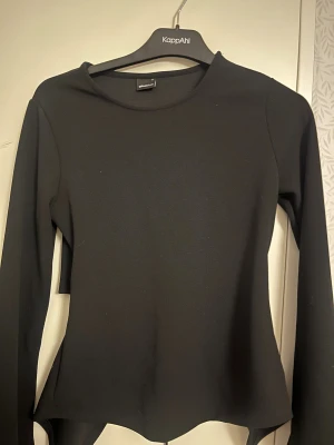 Svart långärmad topp från Gina Tricot - Svart långärmad topp från Gina Tricot med rund halsringning och snygg omlott-detalj baktill som knyts med snörning. Toppens material är stretchigt och har en clean look, perfekt för att lyfta din outfit med en cool twist.